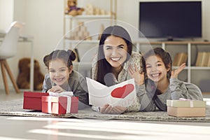 Mother with a postcard in her hands lying on the floor with her daughters among the gifts.