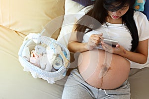 Mother pregnant sitting relax on the bed with diaper and bottle and milk in baby basket, female playing mobile phone