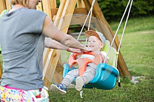 Mother pushing her infant baby boy child on a swing on playground outdoors.