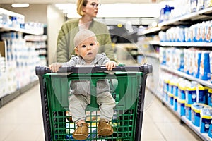 Mother pushing shopping cart with her infant baby boy child down department aisle in supermarket grocery store. Shopping