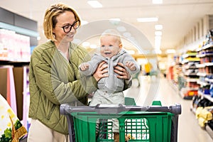 Mother pushing shopping cart with her infant baby boy child down department aisle in supermarket grocery store. Shopping