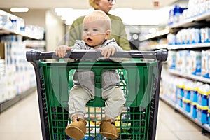 Mother pushing shopping cart with her infant baby boy child down department aisle in supermarket grocery store. Shopping