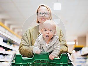 Mother pushing shopping cart with her infant baby boy child down department aisle in supermarket grocery store. Shopping