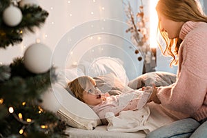 Mother puts her little daughter to bed in a cozy room decorated for christmas.