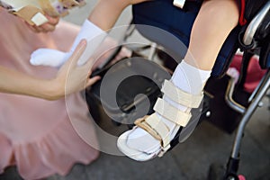 Mother puts on orthosis her daughter legs. Disabled girl sitting on a wheelchair. Child cerebral palsy