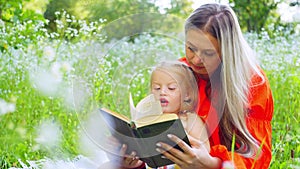 A mother is reading a book to her daughter while sitting on the grass in field.