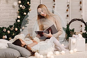 Mother reading book to her little daughter near Christmas tree