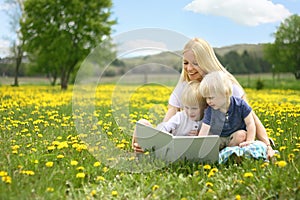 Mother Reading Story Book to Two Young Children Outside in Meadow