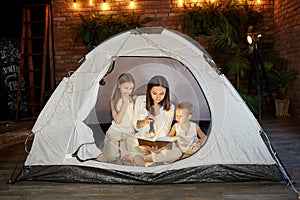 Mother reads a book of fairy tales for her children while sitting in a tent at night. Mom son and daughter reading a book with a