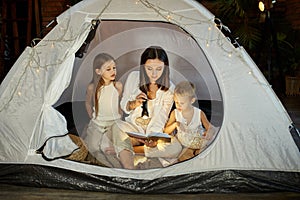 Mother reads a book of fairy tales for her children while sitting in a tent at night. Mom son and daughter reading a book with a