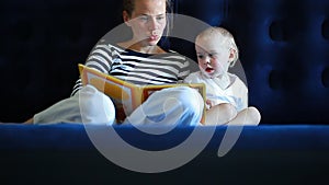 Mother reads a book to a baby sitting on a mattress