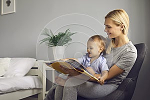 Mother reads a children's book to her little toddler son sitting in a chair in a cozy bedroom.