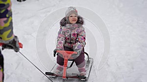 Mother ride a little girl on a sled with a steering wheel on a snow path in the park. Happy child looks around during a