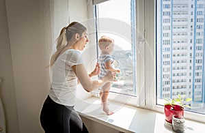 Mother rushing to her baby boy standing on windowsill. Concept of baby in danger