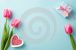 Top view photo of spring flowers pink tulips small giftbox with bow and heart shaped saucer with sprinkles on blue background