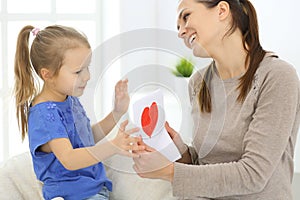 Mother`s day concept. Child daughter congratulates mom and gives her postcard with red heart shape. Mum and girl happy