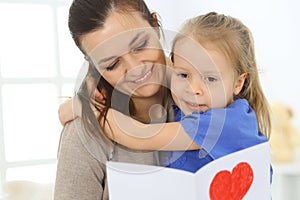 Mother`s day concept. Child daughter congratulates mom and gives her postcard with red heart shape. Mum and girl happy