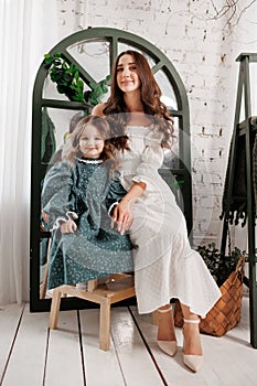 Mother& x27;s day. Elegant young mom and little cute daughter sit and smile, embrace on wooden chair at living room at