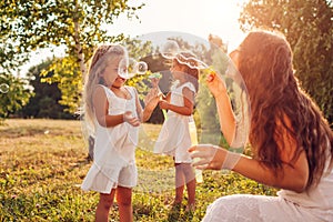 Mother`s day. Woman helps daughters to blow soap bubbles in summer park. Kids having fun playing outdoors