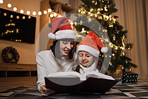 Mother in santa hat reading fairy tale book to her daughter lying on floor near Christmas tree