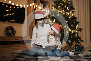 Mother in santa hat reading fairy tale book to her daughter sitting on floor near Christmas tree