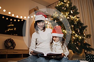 Mother in santa hat reading fairy tale book to her daughter sitting on floor near Christmas tree