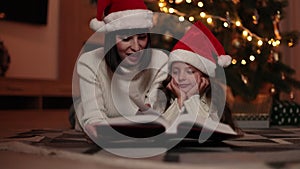 Mother in santa hat reading magical fairy tale book to her daughter lying on floor near Christmas tree