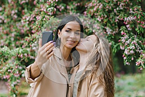 Mother and a schoolgirl daughter take a selfie in the spring in the park. Happy and positive emotions. Daughter kisses mom on the