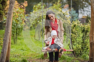 Mother shakes her baby on swing at the autumn park. Happy family spend time together