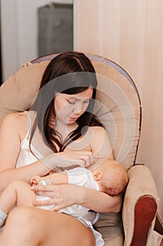 mother in silk nightshirt feeds baby with breast milk in chair. breastfeeding.