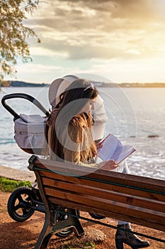 Mother sits on a bench at the sea with her baby and reading a book