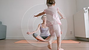 Mother sits in lotus position on yoga mat and little girl runs and plays nearby at home