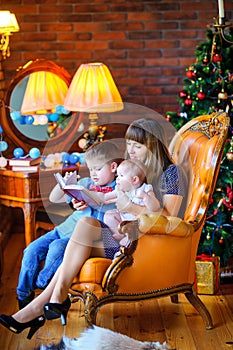 Mother sitting in a chair near the Christmas tree reads tales to lubimim children