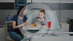 Mother sitting with sick child patient while eating healthy food lunch in hospital ward