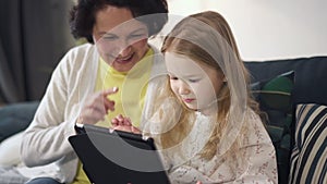 Mother is sitting on sofa, talking and showing to daughter the best using of laptop.