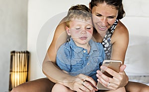 Mother sitting with son on bed using mobile