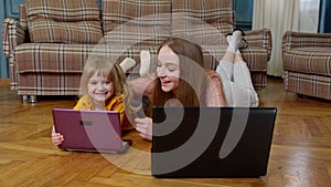 Mother and small daughter child sit on warm wooden floor at home working on laptop, watching cartoon