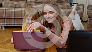 Mother and small daughter child sit on warm wooden floor at home working on laptop, watching cartoon