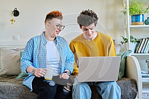 Mother and son 19-20 years old, sitting together on couch at home, looking at laptop screen