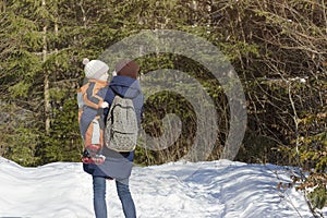 Mother with son in arms and backpack stands against the background of coniferous forest and snow-covered road. Winter day