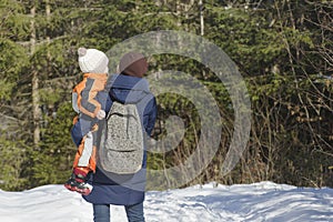 Mother with son in arms and backpack stands against the background of coniferous forest and snow-covered road. Winter day