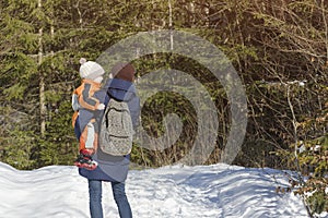 Mother with son in arms and backpack stands against the background of coniferous forest and snow-covered road. Winter day