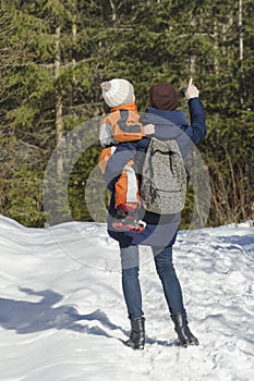 Mother with son in arms and backpack stands against the background of coniferous forest and snow-covered road. Winter day