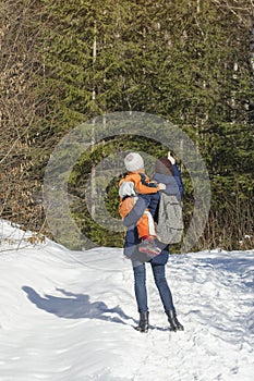 Mother with son in arms and backpack stands against the background of coniferous forest and snow-covered road. Winter day