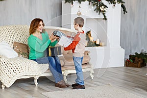 A mother and a son with christmas presents in front of the fur-tree with candles.