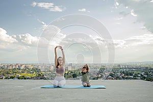 Mother and son doing exercise on the balcony in the background of a city during sunrise or sunset, concept of a healthy