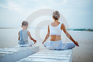 A mother and a son are doing yoga exercises at the seashore of tropic ocean