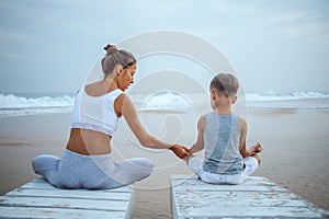 A mother and a son are doing yoga exercises at the seashore of tropic ocean