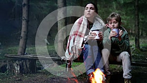 Mother with son drinking the tea and talking near near forest campfire