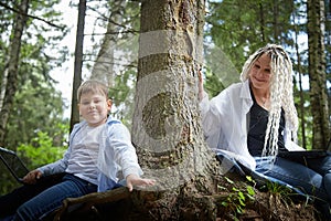 Mother and son with a laptops in forest in summer. Fat young smart teenage boy and woman working with modern IT